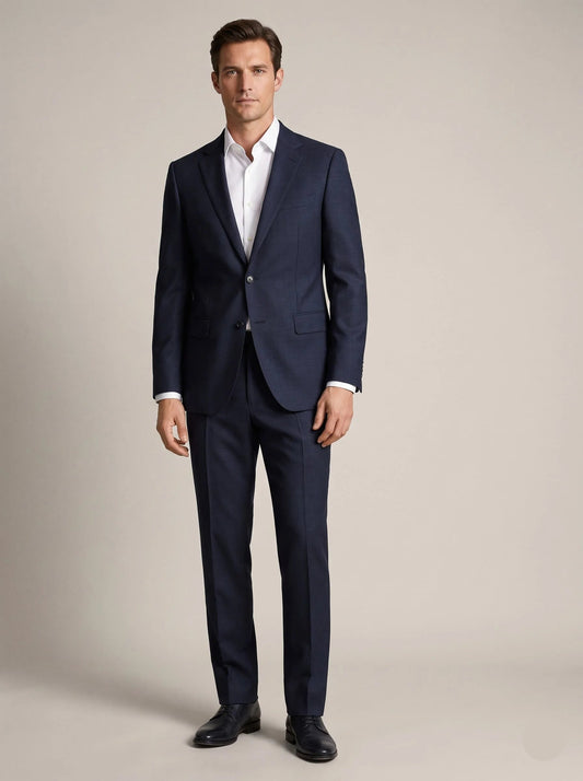 A man modeling a navy blue two-piece suit with a white dress shirt and black dress shoes, standing against a neutral beige studio background.